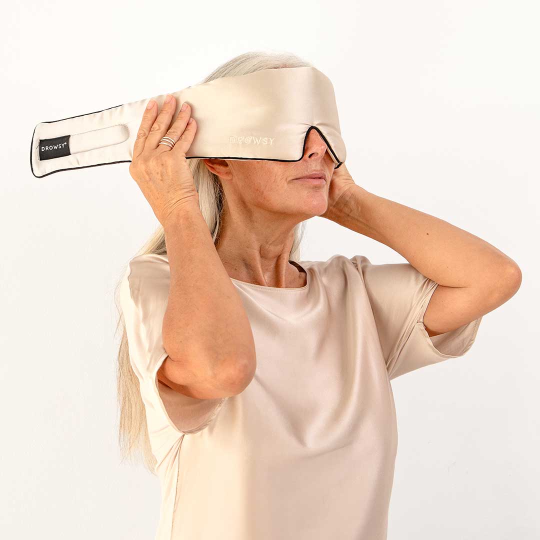 A woman with long gray hair wearing a beige satin top adjusts a large beige silk sleep mask over her eyes against a plain white background.