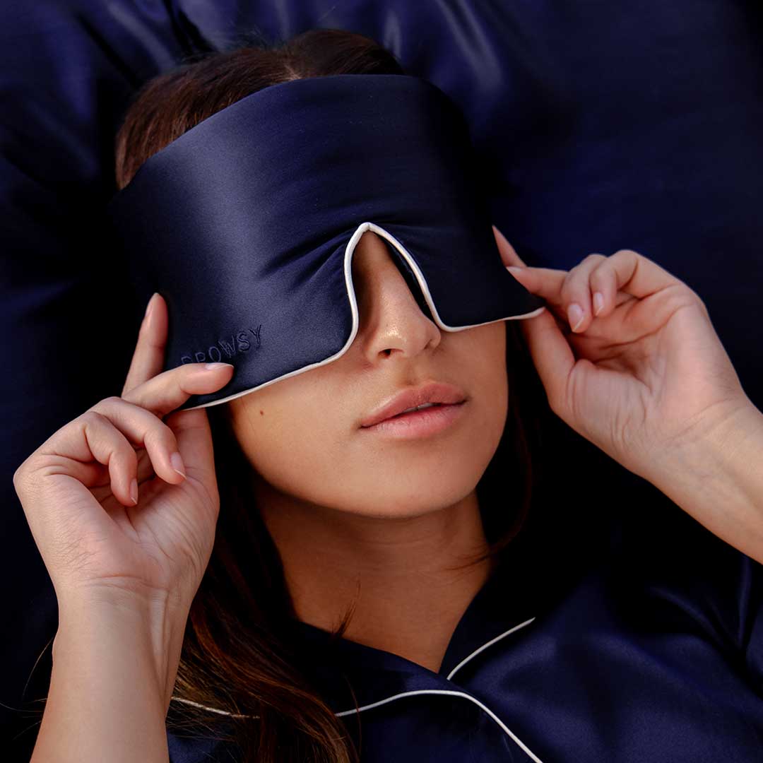 A woman wearing dark blue pajamas is lying down and adjusting a matching dark blue sleep mask over her eyes. The background and bedding are also dark blue.