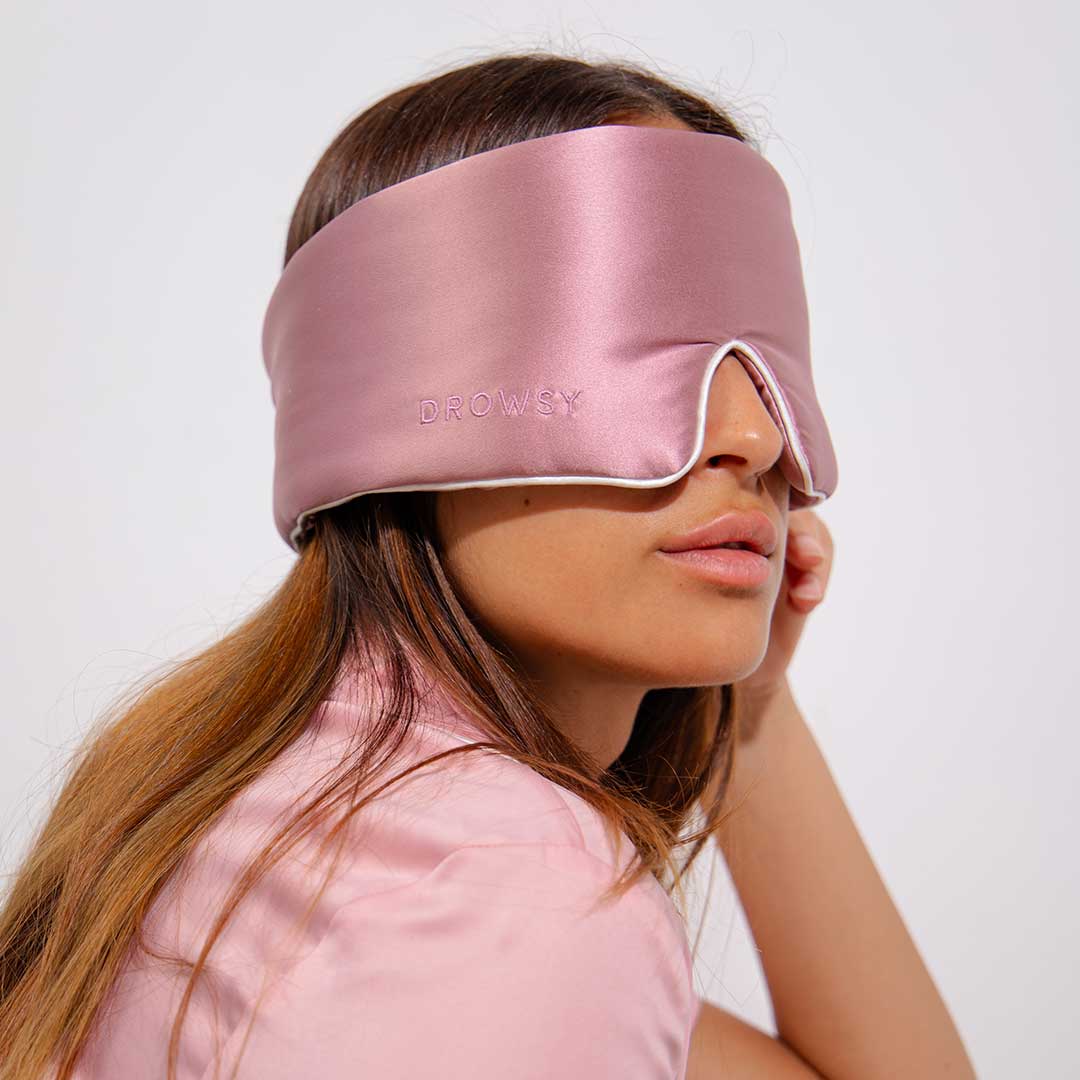 A woman wearing a silky pink sleep mask labeled Drowsy rests her head on her hand. She has long brown hair and is dressed in a matching pink shirt, with a neutral background behind her.