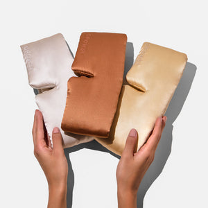 Two hands hold the Drowsy Besties Pack: three luxury mulberry silk sleep masks in beige, copper, and gold on a white background. Each mask is embroidered with “DROWSY,” offering the ultimate in beauty sleep.