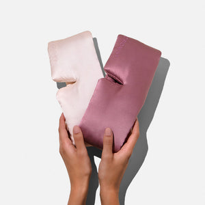 Two hands hold two soft, silky, L-shaped mulberry silk cushions—one light pink, one mauve—from the Lovers Pack by Drowsy, perfect for beauty sleep, shown against a plain white background.