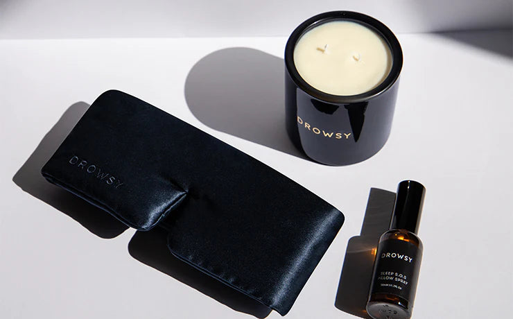 A black silk sleep mask, a black candle, and a small spray bottle labeled Sleep S.O.S. Pillow Spray, all branded Drowsy, are arranged on a white surface with soft shadows.