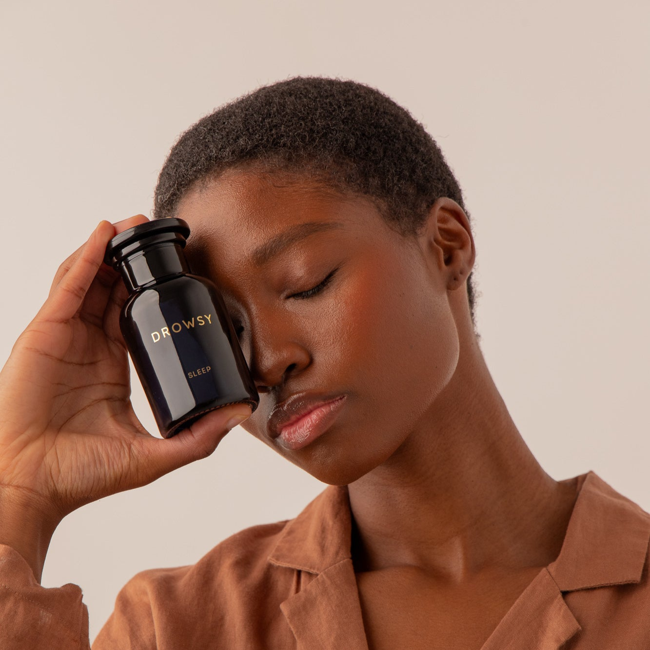 A person with short hair and closed eyes holds a black drowsysleepco Supplements - Sleep bottle to their face, exuding relaxation in a brown top against a neutral background—capturing the calm before restful sleep.
