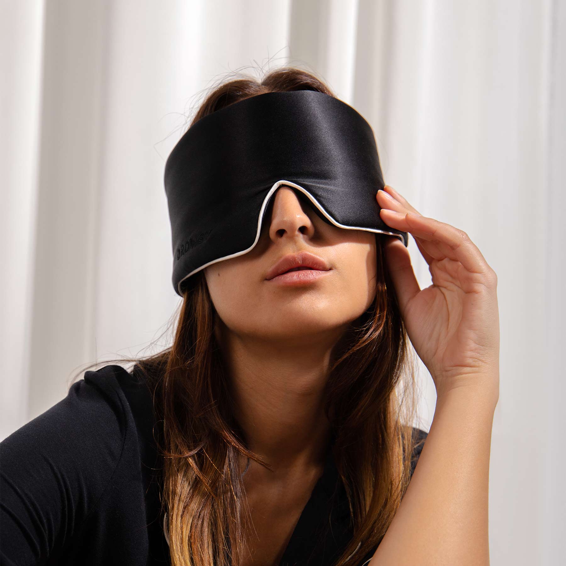 A woman with long brown hair adjusts the head strap of the us.drowsysleepco Black Jade - Pipe Dreams Edition silk sleep mask while seated in front of white curtains.