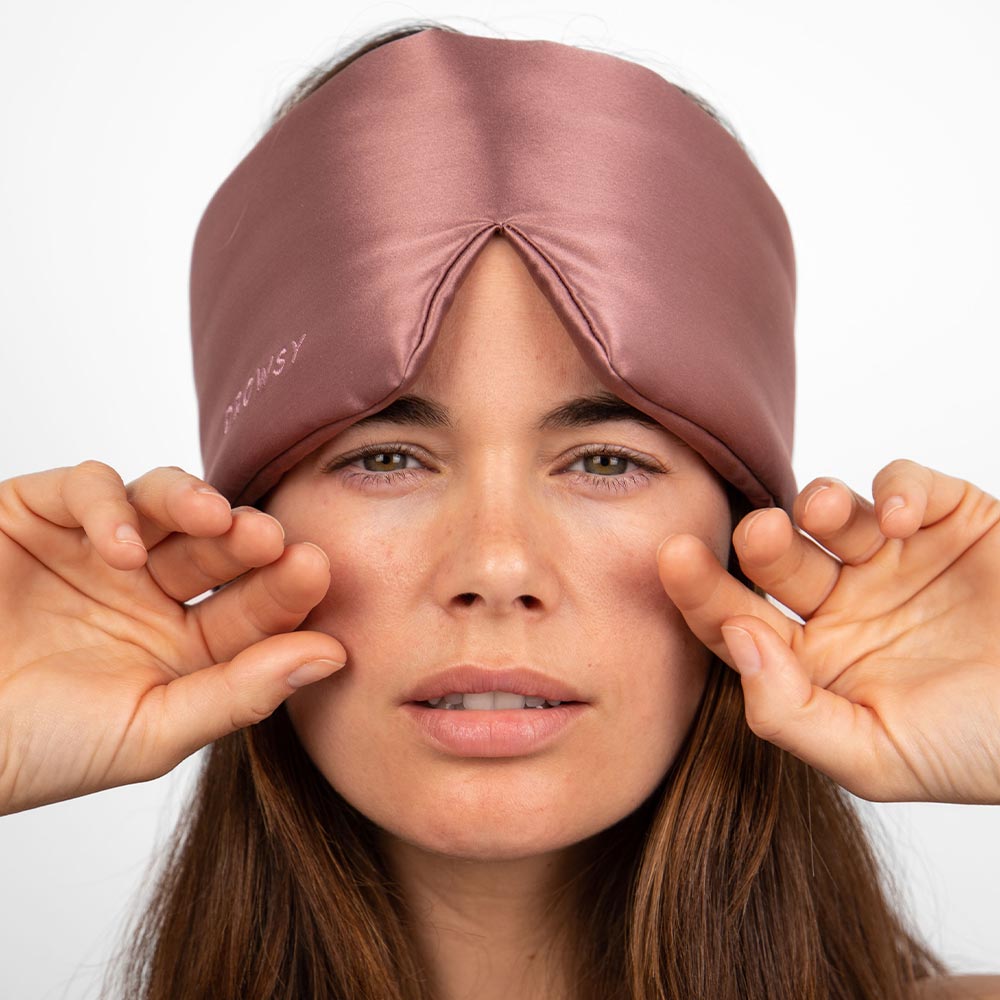 A woman wearing the drowsysleepco Sensual Sleep Bundle - Damask Rose mask on her forehead gently touches it with both hands, looking at the camera with a calm expression that evokes the soothing luxury of silk.