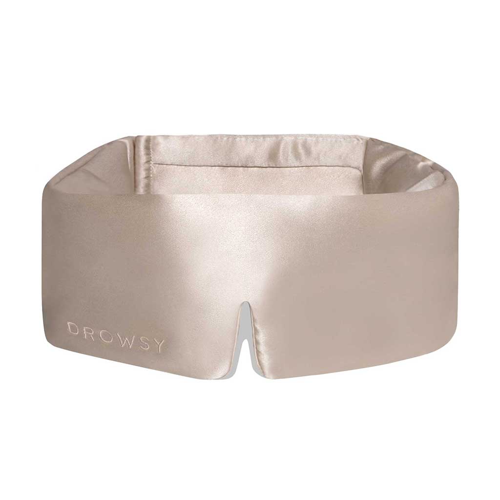 The Dusty Gold eye mask by us.drowsysleepco is a beige silk mask with DROWSY in small letters on the left, featuring a wide, cushioned blackout design and a small notch at the bottom center.