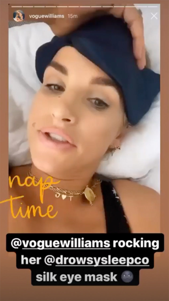 A woman lies on a bed wearing a navy silk eye mask pushed up on her forehead. Text on the image reads nap time and @voguewilliams rocking her @drowsysleepco silk eye mask.