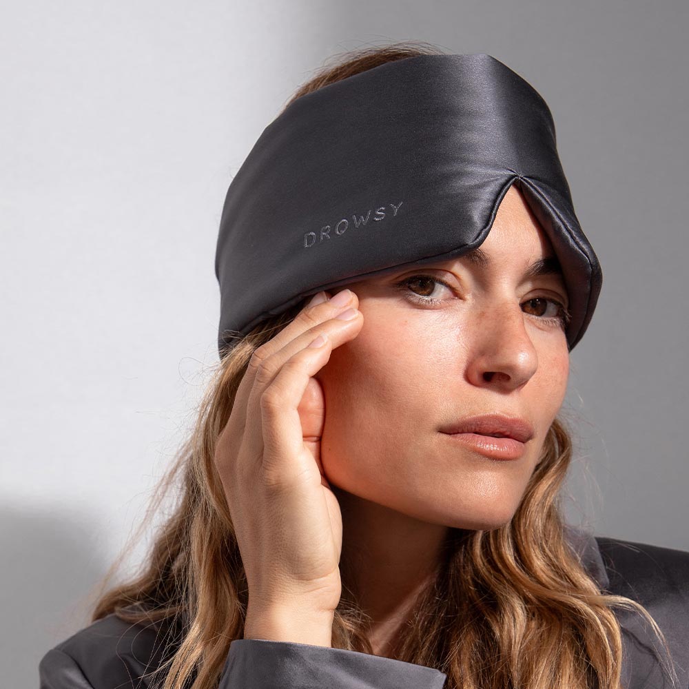 A woman wearing a dark silk sleep mask labeled DROWSY gently touches her face, looking relaxed against a light gray background.