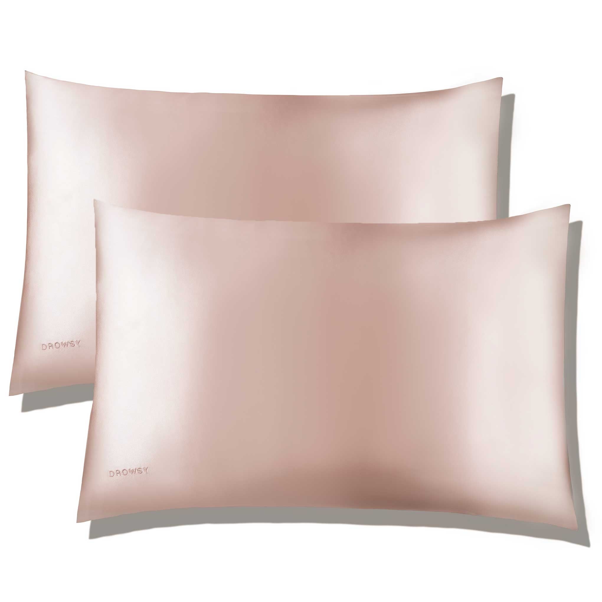 Two Sunset Pink Silk Pillowcases from us.drowsysleepco, each with DROWZY embroidered in the corner, are stacked slightly askew. Their subtle sheen highlights these silk essentials for hair and skin care against a plain white background.