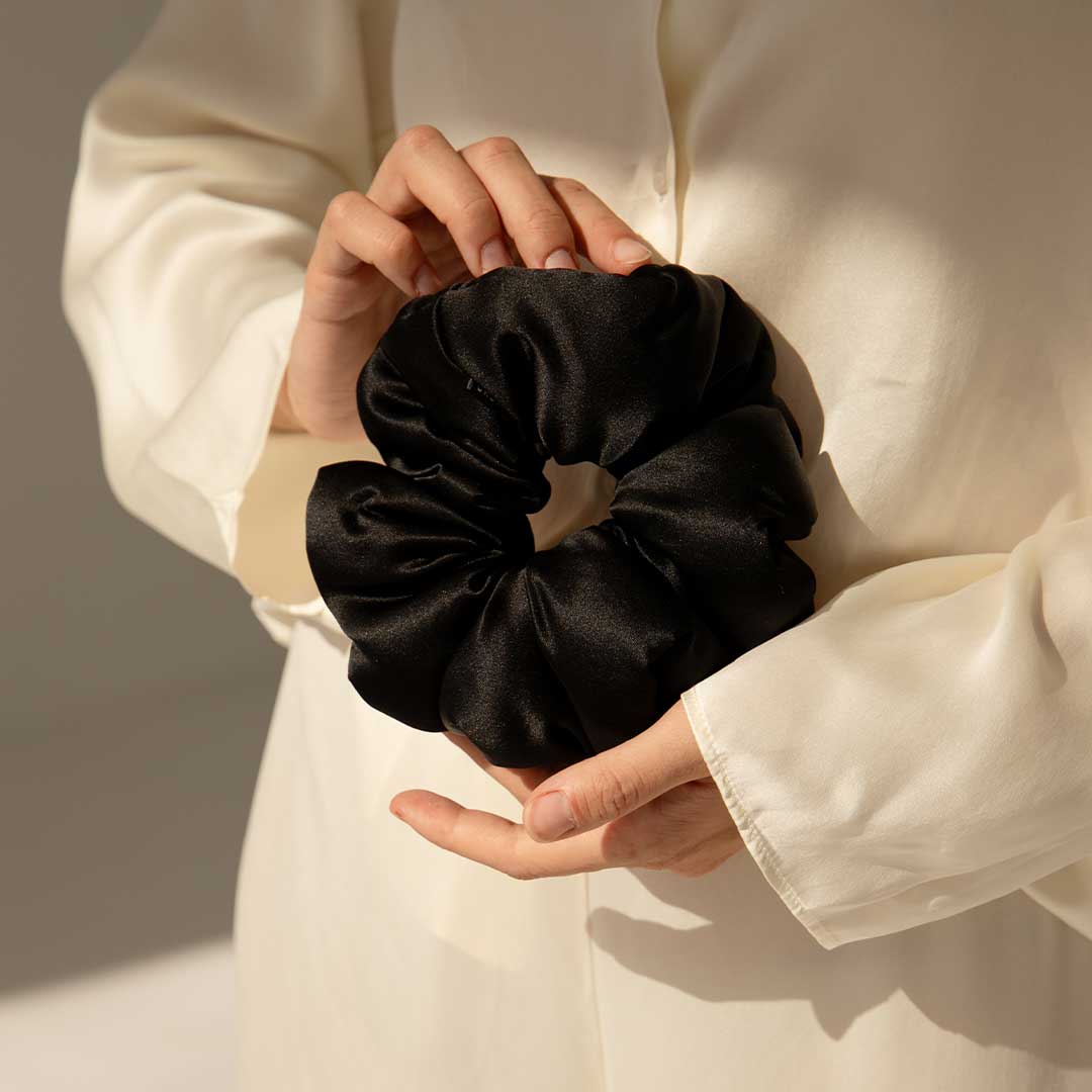 A person wearing a cream-colored, long-sleeve shirt gently holds a large black satin scrunchie with both hands against a neutral background.
