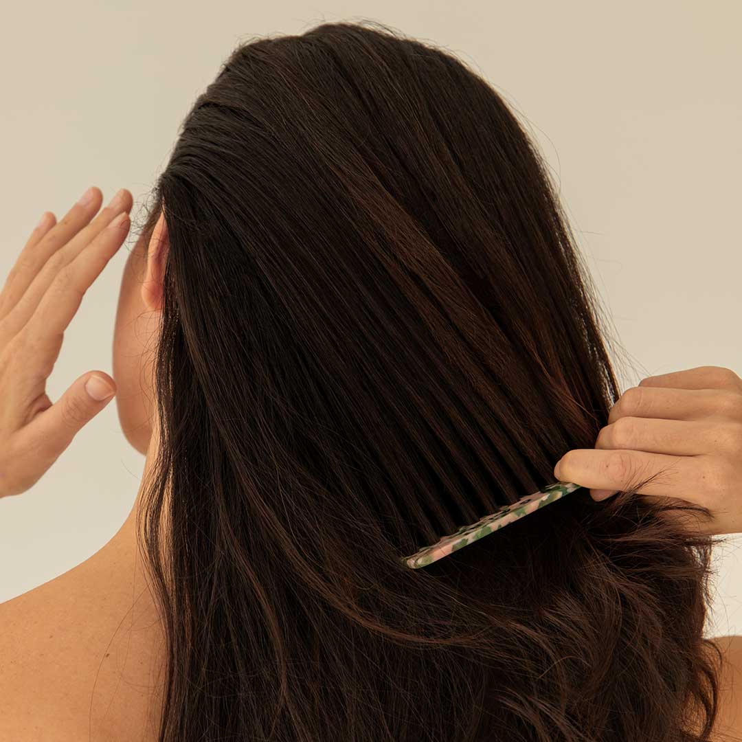 A person with long, dark hair seen from behind is combing their hair with a patterned comb while raising their other hand near their head against a plain background.