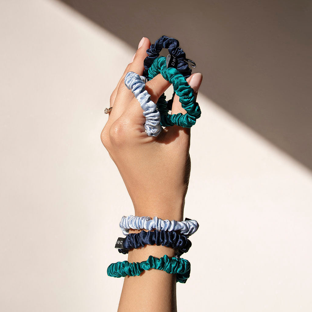 A hand holds several scrunchies in shades of green, navy blue, and light blue, with more scrunchies worn on the wrist and fingers, against a neutral, sunlit background.