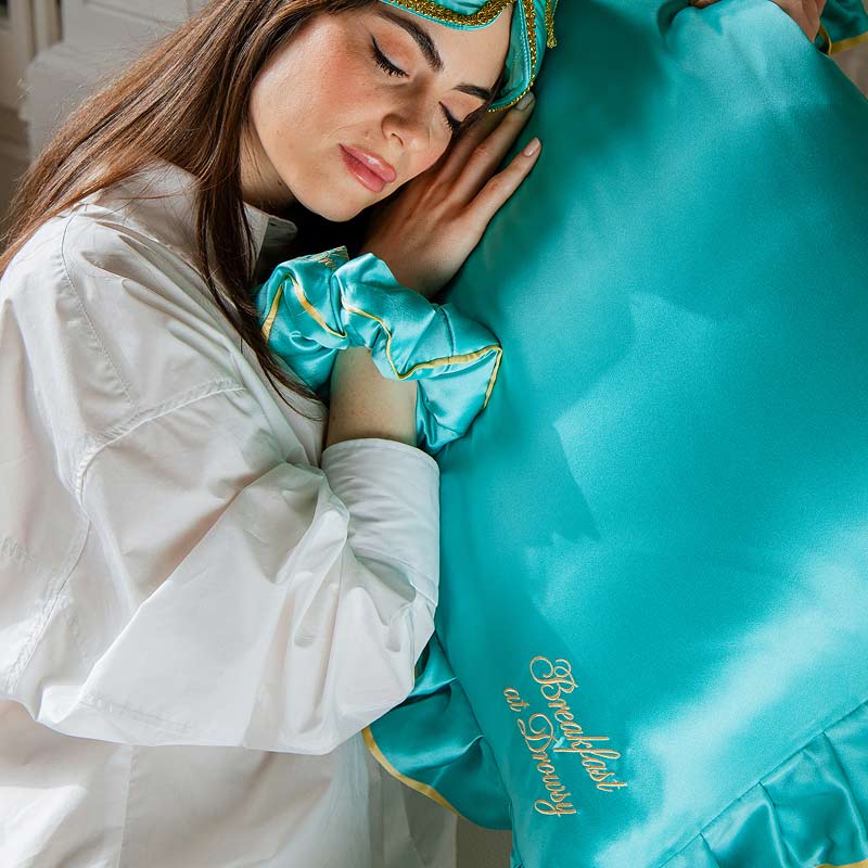 A woman in a white shirt, turquoise silk sleep mask, and matching scrunchie from the drowsysleepco Hollywood Experience bundle - Original rests on a large turquoise satin pillow with gold trim and "Beachlash et Dreamy" embroidery.