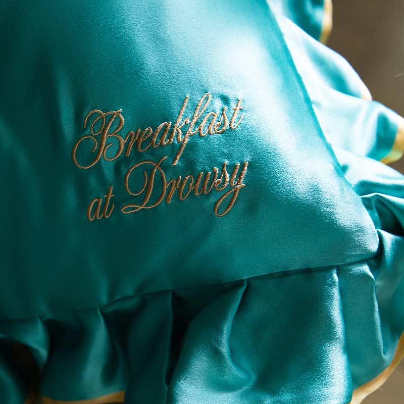 A close-up of the drowsysleepco Breakfast at Drowsy - Turquoise Pillowcase, a teal satin pillow with ruffled edges and gold embroidered text—an elegant, hair-protecting and anti-sleep crease silk pillowcase.