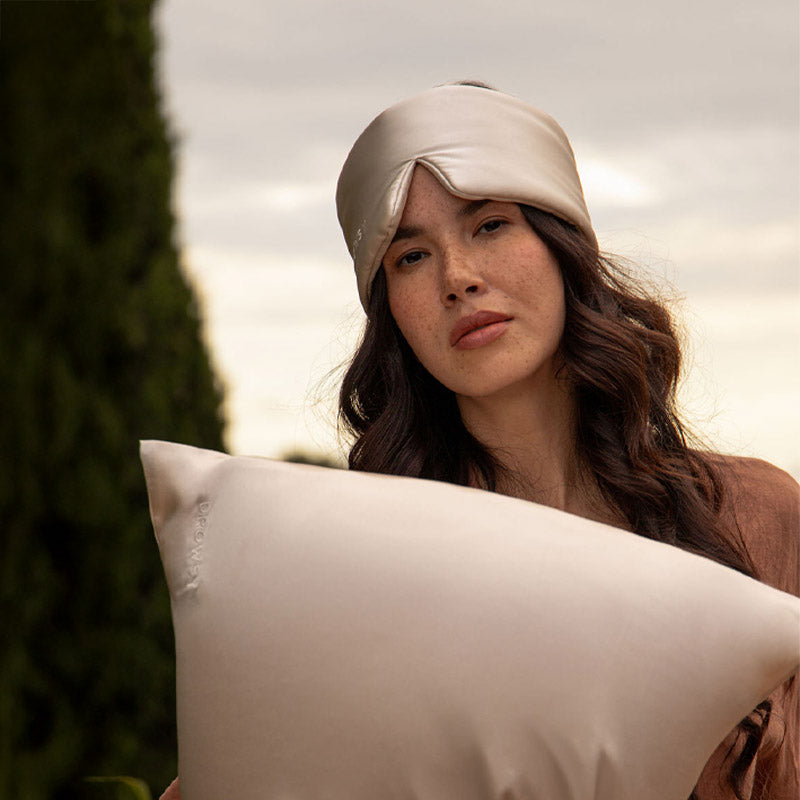 A woman with long dark hair wears the drowsysleepco Sleep Week Ritual beige silk sleep mask on her forehead and holds a matching pillowcase, standing outdoors amid greenery and beneath a cloudy sky.