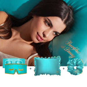 A woman with dark hair relaxes on a turquoise satin pillow embroidered with “Breakfast at Drusy,” alongside the Hollywood Experience Bundle by Drowsy: a turquoise sleep mask, pillowcase, and scrunchie with gold accents for beauty sleep.