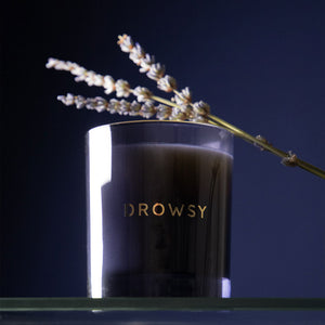 The Sleep Ritual Candle 220g by drowsysleepco stands on a glass surface with lavender on top, its sleep-inducing aroma filling the air. The candle features “Drowsy” in gold lettering, set against a deep blue backdrop.