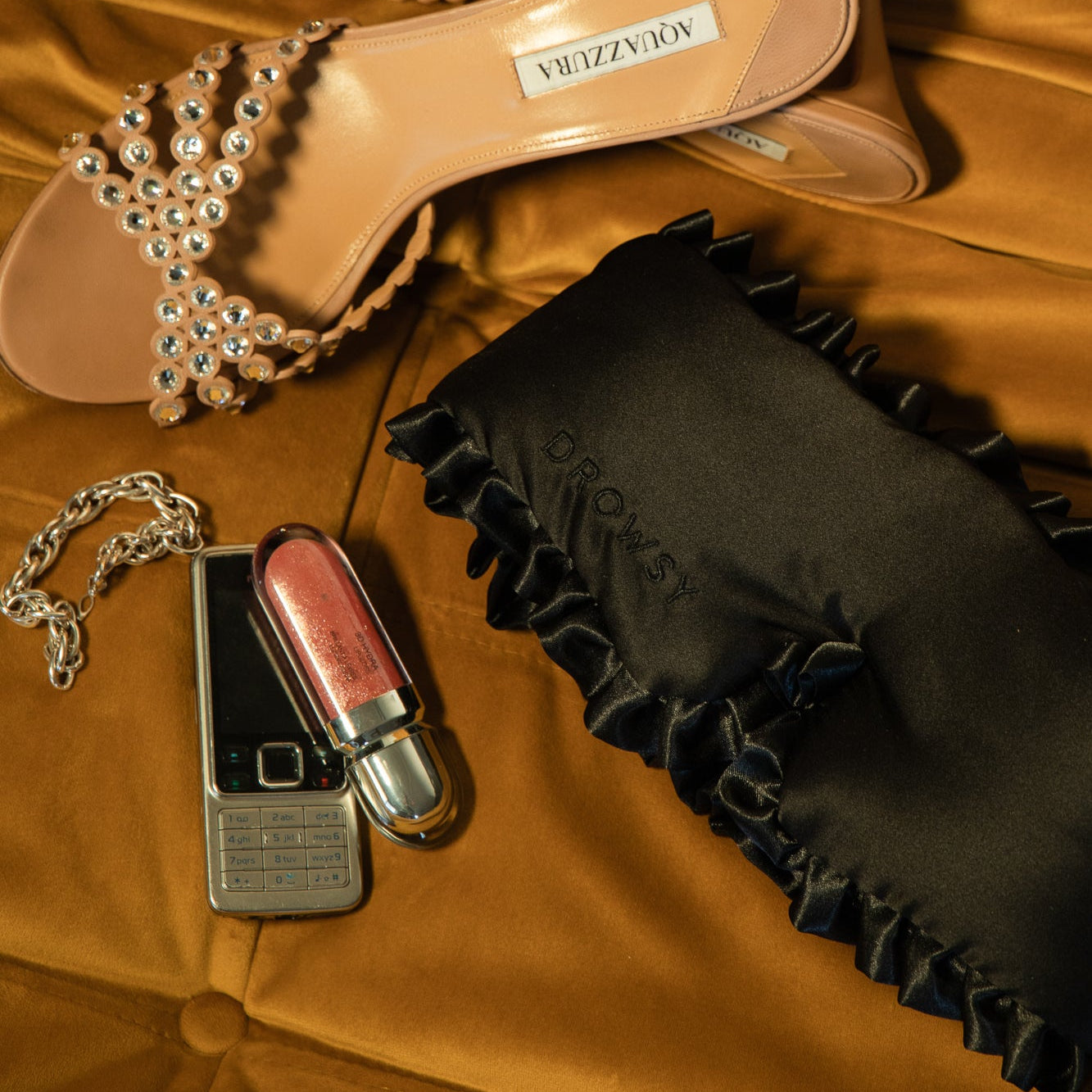 A jeweled sandal, Drowsy’s Sleep and the City black satin sleep mask, a silver flip phone, and shiny pink lipstick with a chain—all arranged on gold velvet.