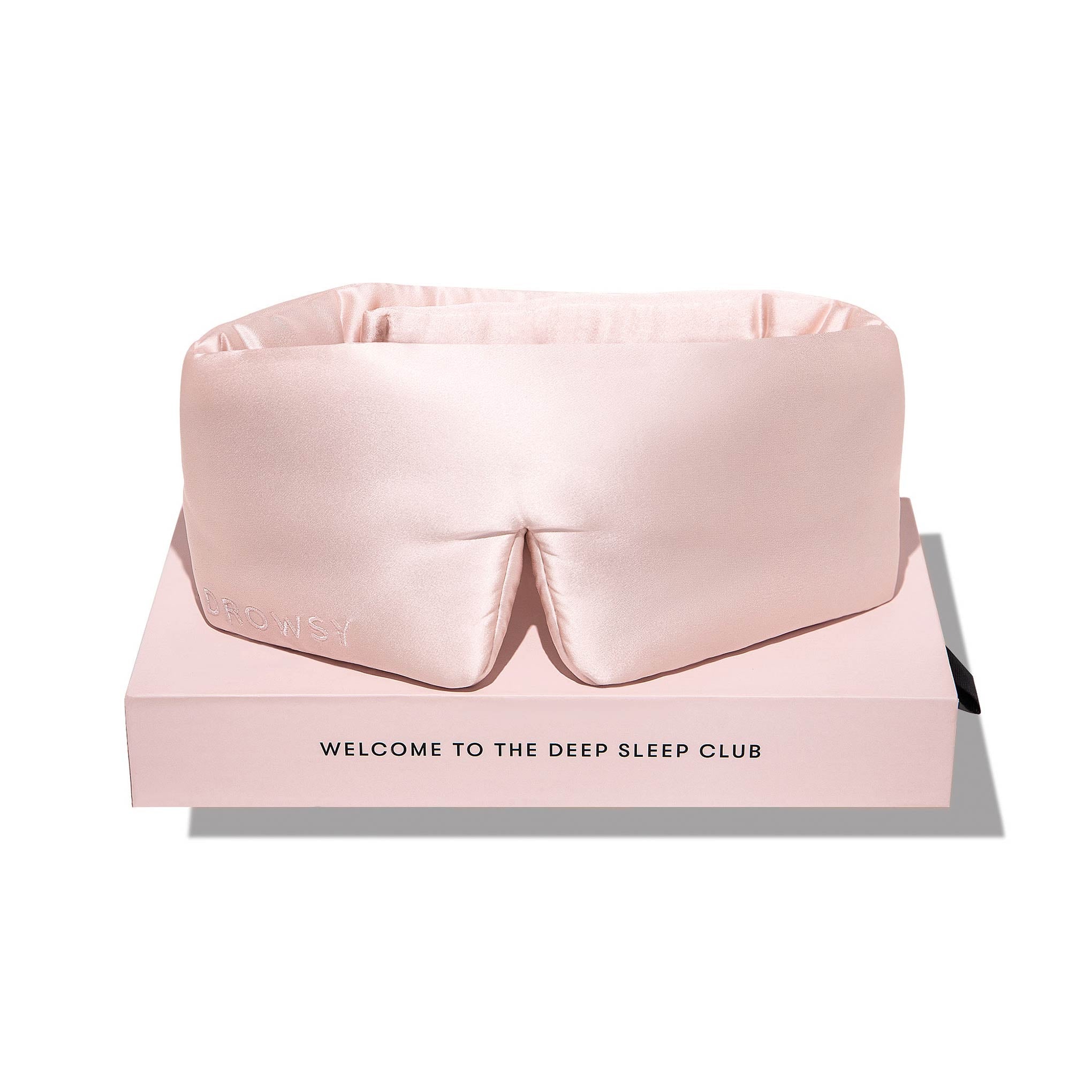 The Sunset Pink silk eye mask by us.drowsysleepco rests on a matching box labeled “WELCOME TO THE DEEP SLEEP CLUB.” This plush mask, with a discreet logo, elevates your nighttime routine with a luxurious touch.