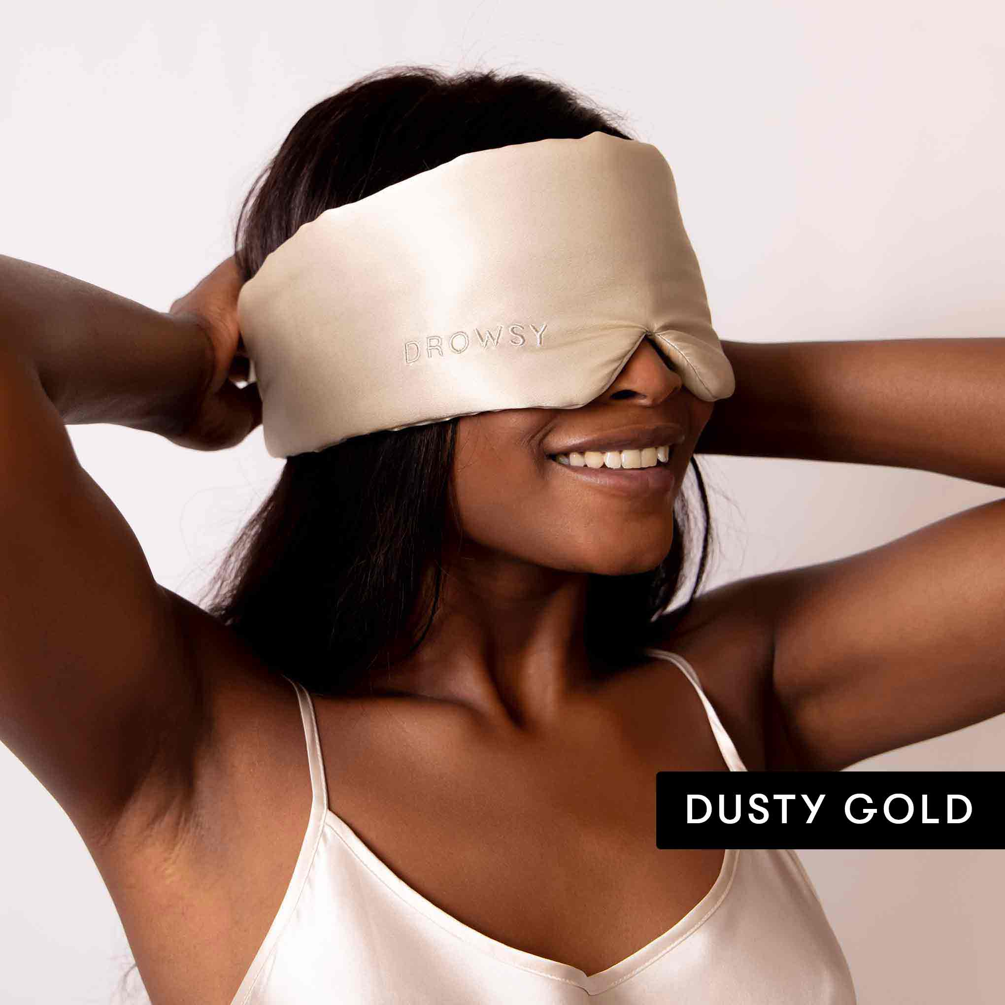 A smiling woman lifts a luxury silk Drowsy mask over her eyes against a white background, ready for beauty sleep. DUSTY GOLD appears in the corner. The Lovers Pack by Drowsy is featured.