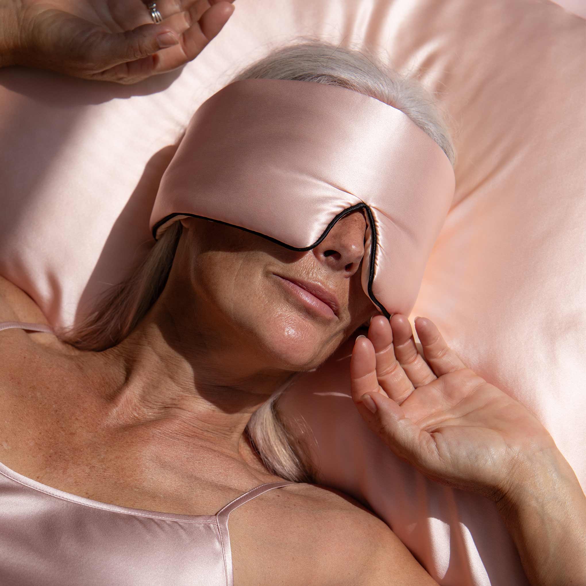 A woman with gray hair lies on a bed, softly smiling as she enjoys her beauty sleep wearing the us.drowsysleepco Sunset Pink – Pipe Dreams Edition silk sleep mask with adjustable strap and matching satin pillowcase.