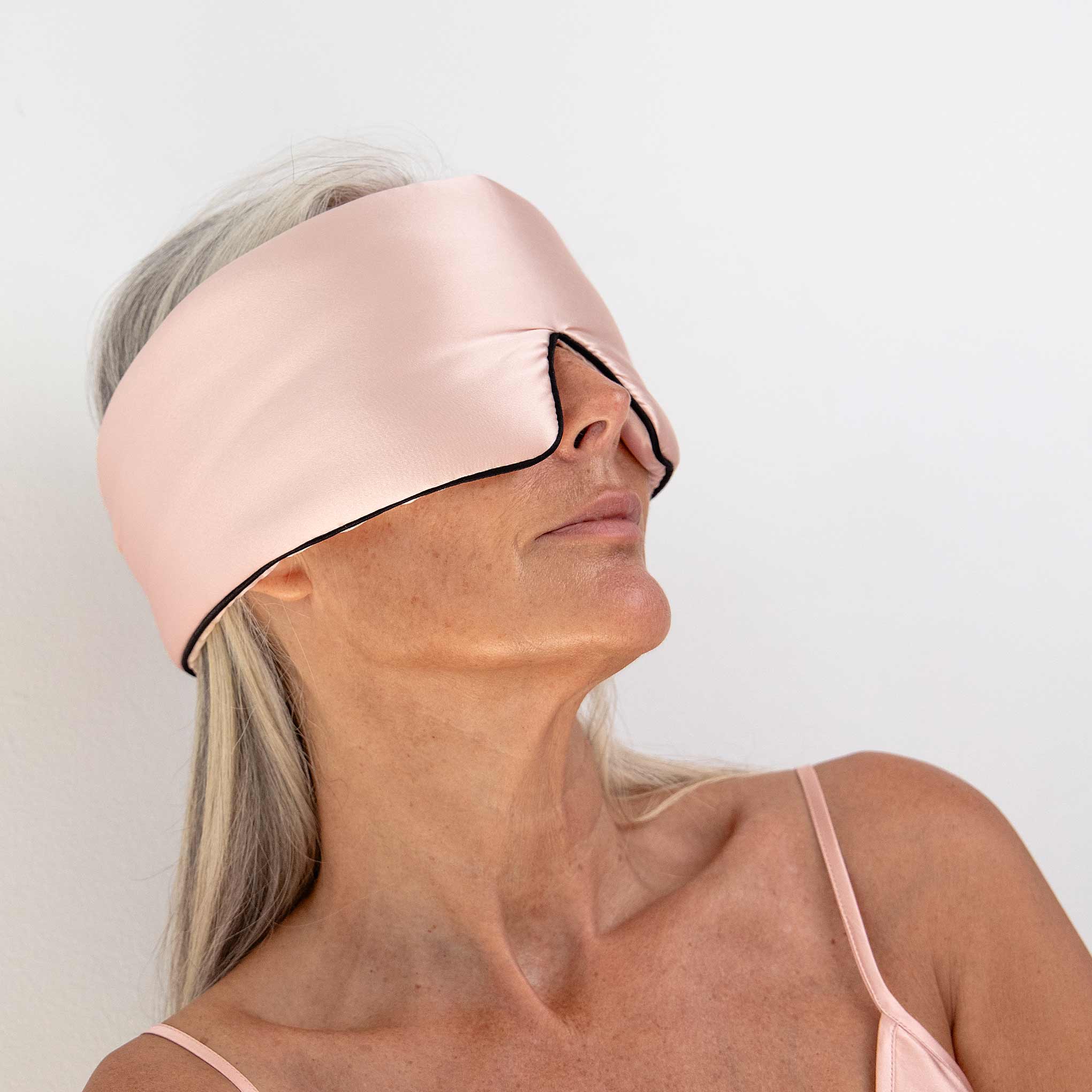 A woman with long gray hair stands against a white background, wearing us.drowsysleepcos Sunset Pink - Pipe Dreams Edition silk sleep mask with an adjustable strap and a pink camisole, ready for restful beauty sleep.