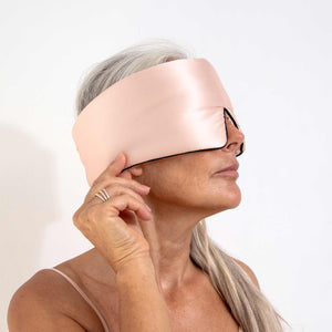 A woman with long gray hair adjusts the us.drowsysleepco Sunset Pink - Pipe Dreams Edition silk sleep mask, featuring an adjustable strap, as she faces sideways against a plain white background, ready for restful beauty sleep.