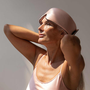 A woman with long gray hair, wearing the Sunset Pink - Pipe Dreams Edition sleepwear by us.drowsysleepco, looks upward with her hands behind her head, basking in soft natural light as if reveling in beauty sleep.