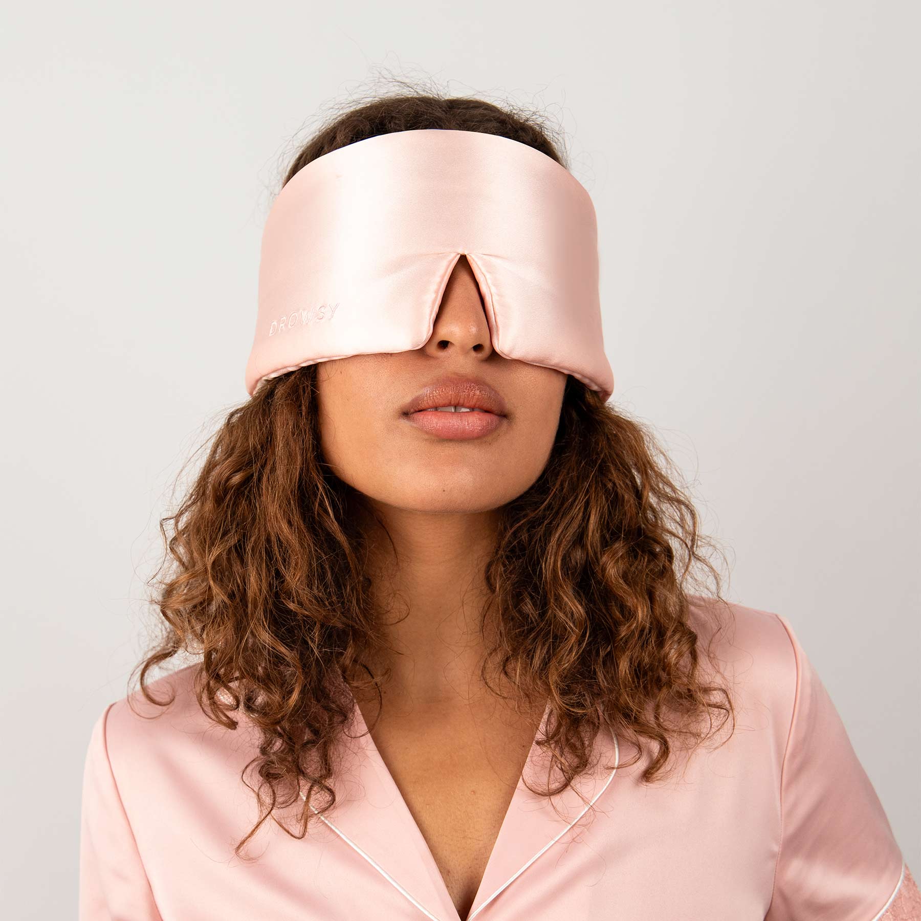 A woman with curly hair stands against a white background, wearing the us.drowsysleepco Sunset Pink silk eye mask and matching pajamas—ready for beauty sleep.