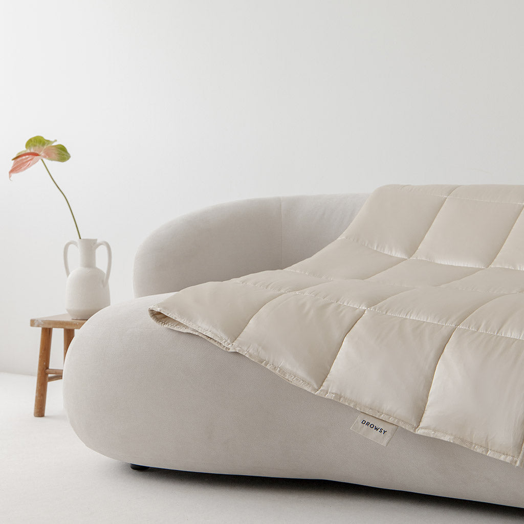 The Dusty Gold Silk Weighted Blanket by us.drowsysleepco rests on a curved, light sofa. Nearby, a wooden side table with a white vase and plant adds to the minimalist décor, promising deep relaxation and an unmatched sleep experience.