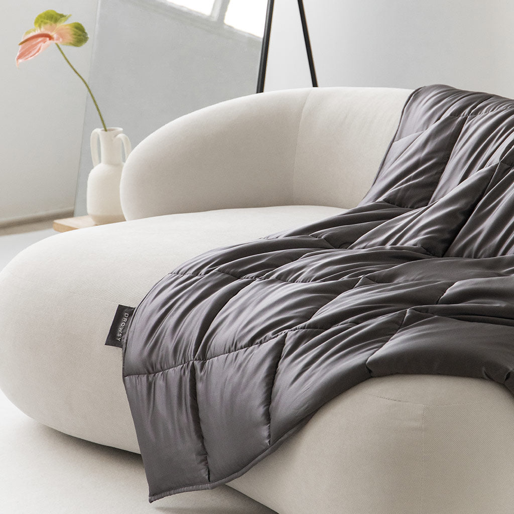 A soft, light sofa is draped with the us.drowsysleepco Moonlight Shadow Silk Weighted Blanket in a modern, minimalistic room. In the background, a white vase with a pink flower on a wooden table adds to the serene atmosphere.
