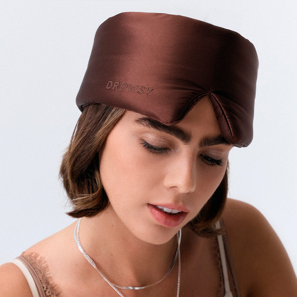 A woman with short brown hair wears a dark brown silk sleep mask labeled 