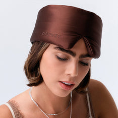 A woman with short brown hair wears a dark brown silk sleep mask labeled 