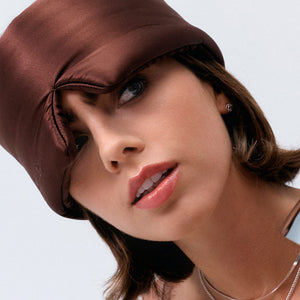 A woman with short brown hair wears the drowsysleepco Cocoa Glow sleep mask, styled chicly over one eye like a shiny brown hat. She sports light makeup, pink lipstick, small earrings, and a silver necklace against a light background.