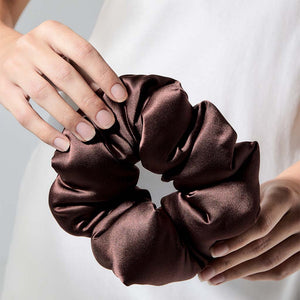A person in a white top holds the us.drowsysleepco Cocoa Glow - Padded Scrunchie, an ideal hair sleep accessory, with both hands against a neutral background.