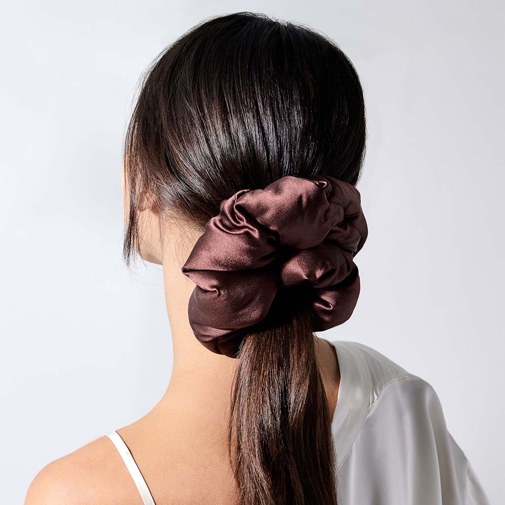 A woman with straight, dark brown hair secures her low ponytail with the us.drowsysleepco Cocoa Glow - Padded Scrunchie, an ideal hair sleep accessory. She wears a white thin-strapped top with an off-the-shoulder sleeve.