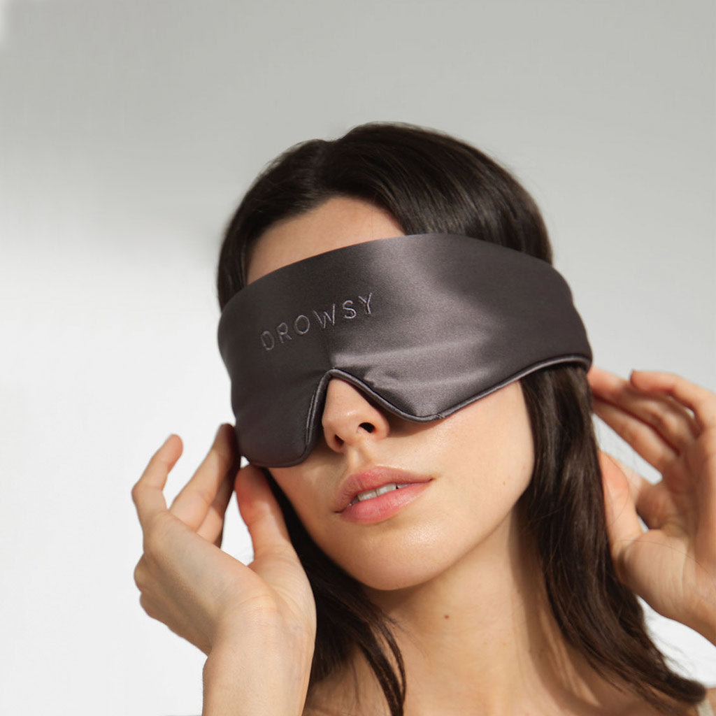 A woman adjusts the Featherweight Silk Eye Mask Moonlight Shadow from us.drowsysleepco over her eyes, gently pressing the smooth, dark gray silk, total black-out mask against a neutral background.