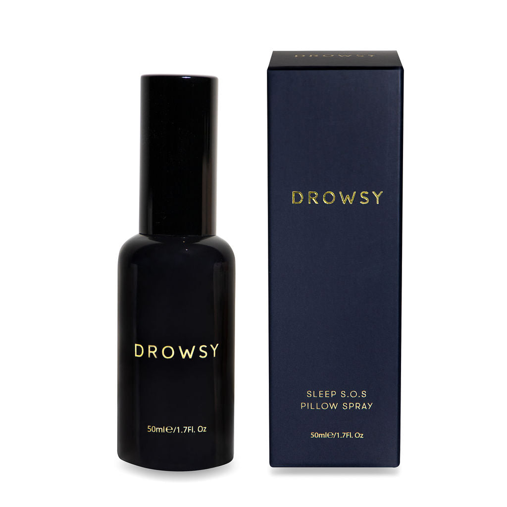 A dark bottle labeled Drowsy stands next to its matching box, which reads Drowsy Sleep S.O.S Pillow Spray, 50ml/1.7fl oz in gold text on a dark background.