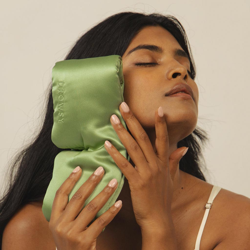 A woman with long dark hair holds a Pistachio Green silk eye mask from us.drowsysleepco against her face, eyes closed in serene relaxation, as if embracing beauty sleep, set against a neutral background.