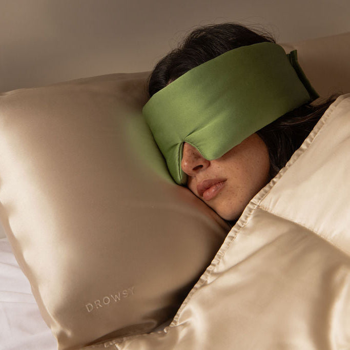 A woman enjoys her beauty sleep with drowsysleepco's Sleep Week Ritual: a beige satin pillowcase embroidered with DROWSY, a matching blanket, and a green silk sleep mask.