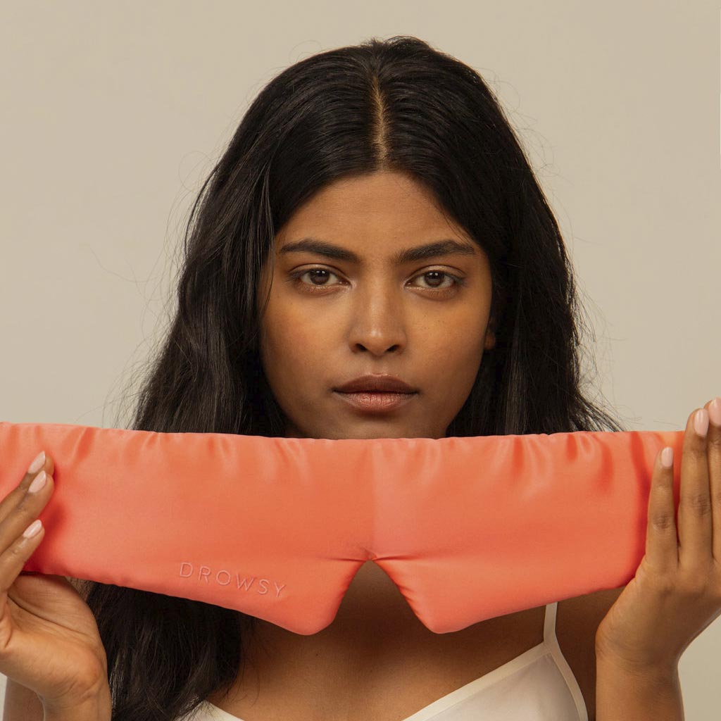 A woman with long dark hair holds the Summer Coral silk eye mask by us.drowsysleepco in front of her chest, looking directly at the camera against a plain beige background—ready for ultimate beauty sleep.