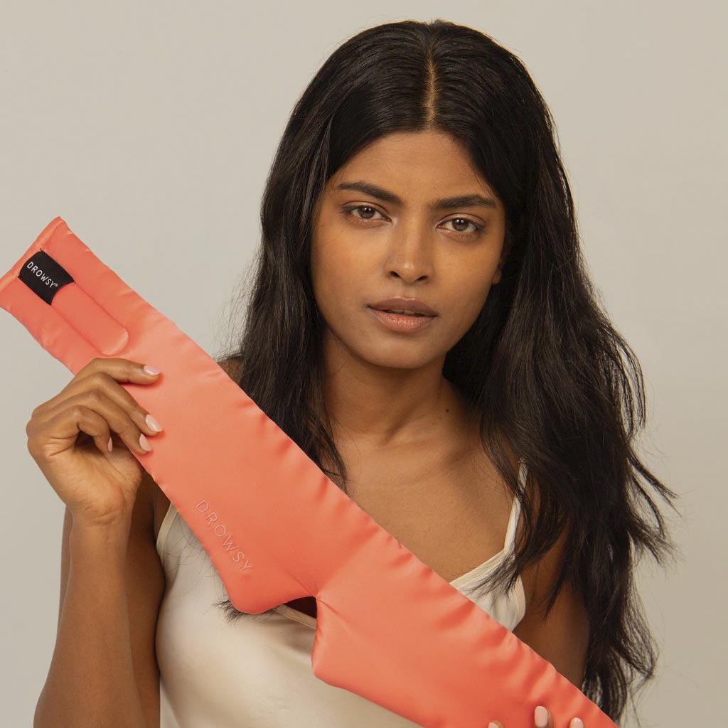 A woman with long dark hair and a neutral expression holds a us.drowsysleepco Summer Coral silk eye mask, perfect for beauty sleep, while wearing a white tank top against a plain beige background.