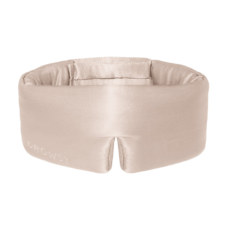 The Dusty Gold eye mask by drowsysleepco is a beige silk mask with DROWSY in small letters on the left, featuring a wide, cushioned blackout design and a small notch at the bottom center.