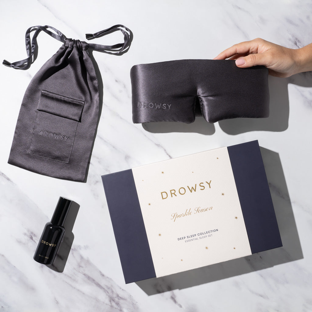 A hand holds us.drowsysleepco’s Moonlight Shadow - Deep Sleep Collection: a luxury silk sleep mask with matching pouch, deep sleep pillow spray in black bottle, and a white gift box with gold stars—perfect for restful nights.
