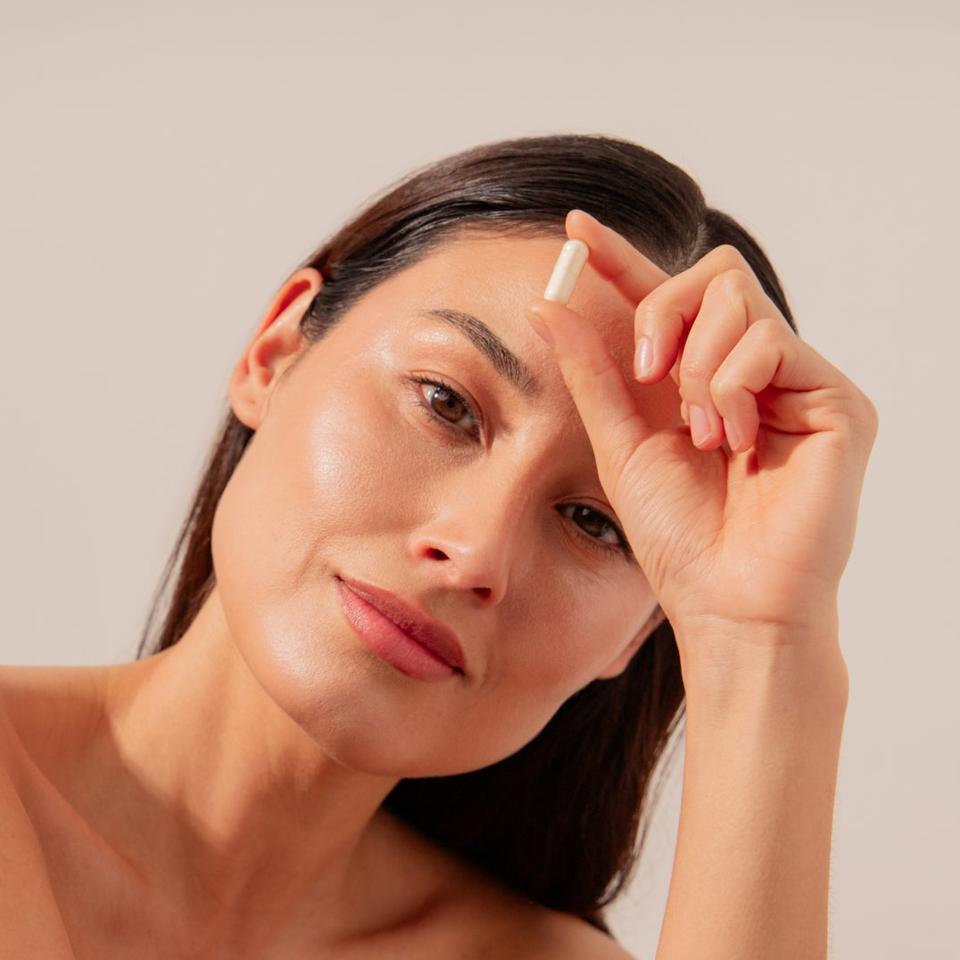 A woman with straight dark hair holds a white capsule of drowsysleepco Supplements - Beauty near her face, suggesting its a skin supplement for a youthful complexion, as she looks at the camera against a plain light background.