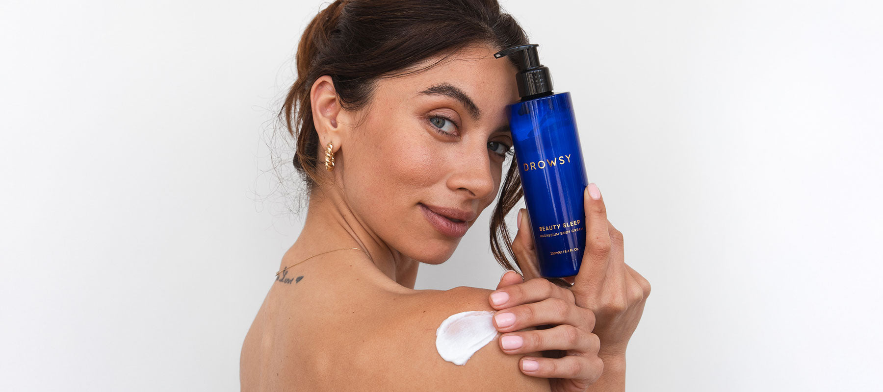 A woman with light brown hair holds a blue bottle labeled Drowsy near her face. She smiles slightly and has lotion applied to her shoulder. The background is plain white.