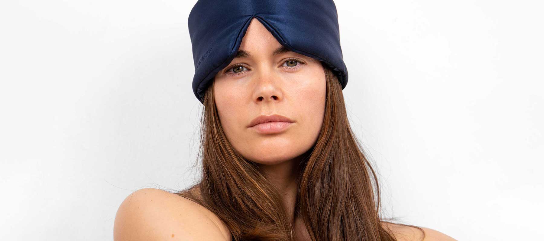 A woman with long brown hair wears a dark blue satin sleep mask pushed up on her forehead, exposing her face. She is looking straight at the camera with a neutral expression against a plain white background.