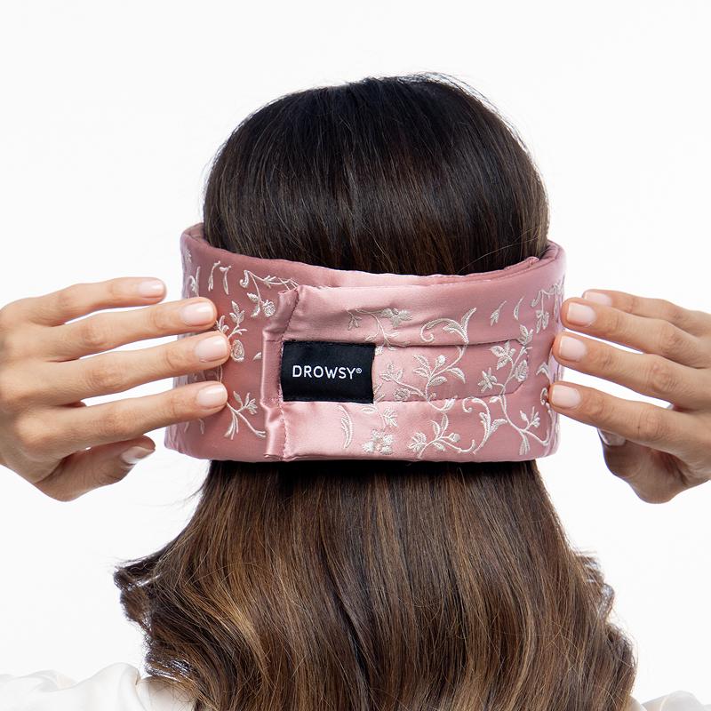 A person with brown hair putting on a dusty pink Pride & Prejudice - English Rose Sleep Mask, decorated with a white floral pattern and a central us.drowsysleepco brand tag.