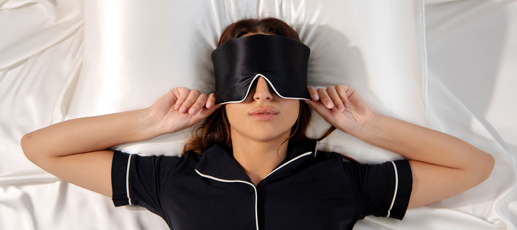 A woman lying on a bed with white sheets wears a black sleep mask and matching black pajamas, holding the mask in place over her eyes with both hands.