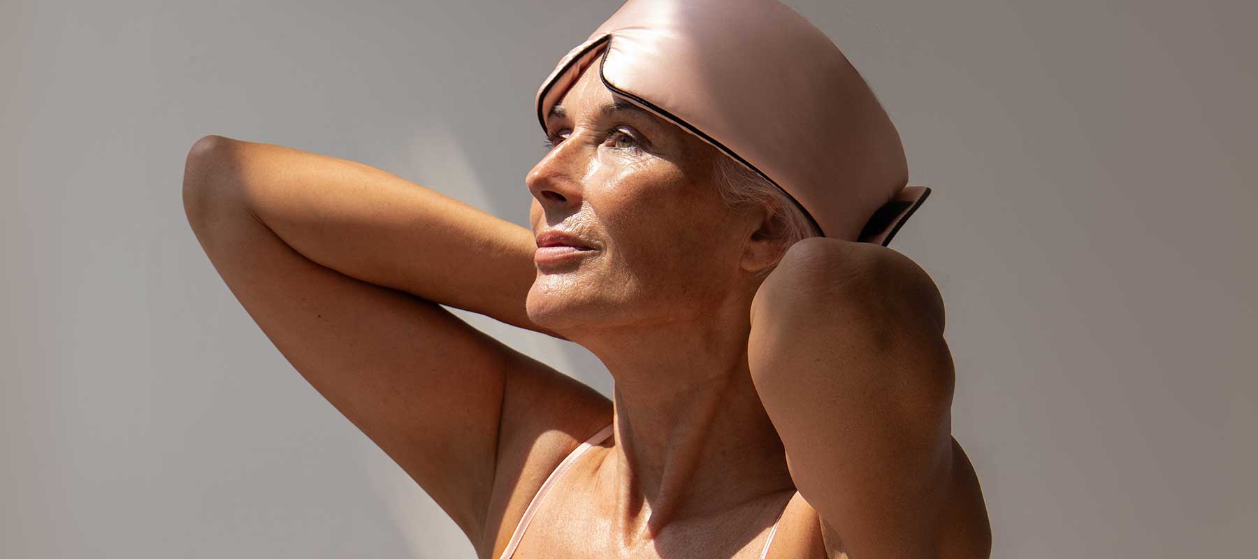 A woman with gray hair, wearing a pink sleep mask on her head and a light tank top, looks up with her hands behind her neck against a plain background, bathed in natural light.